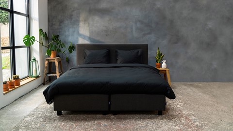 Een bed met zwarte lakens en kussens in een kamer met een grijze muur