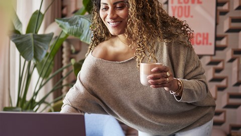 Een vrouw die een kopje vasthoudt en naar een laptop kijkt