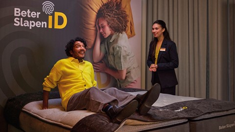 een man in gele trui zittend op een bed met een vrouw die achter hem staat